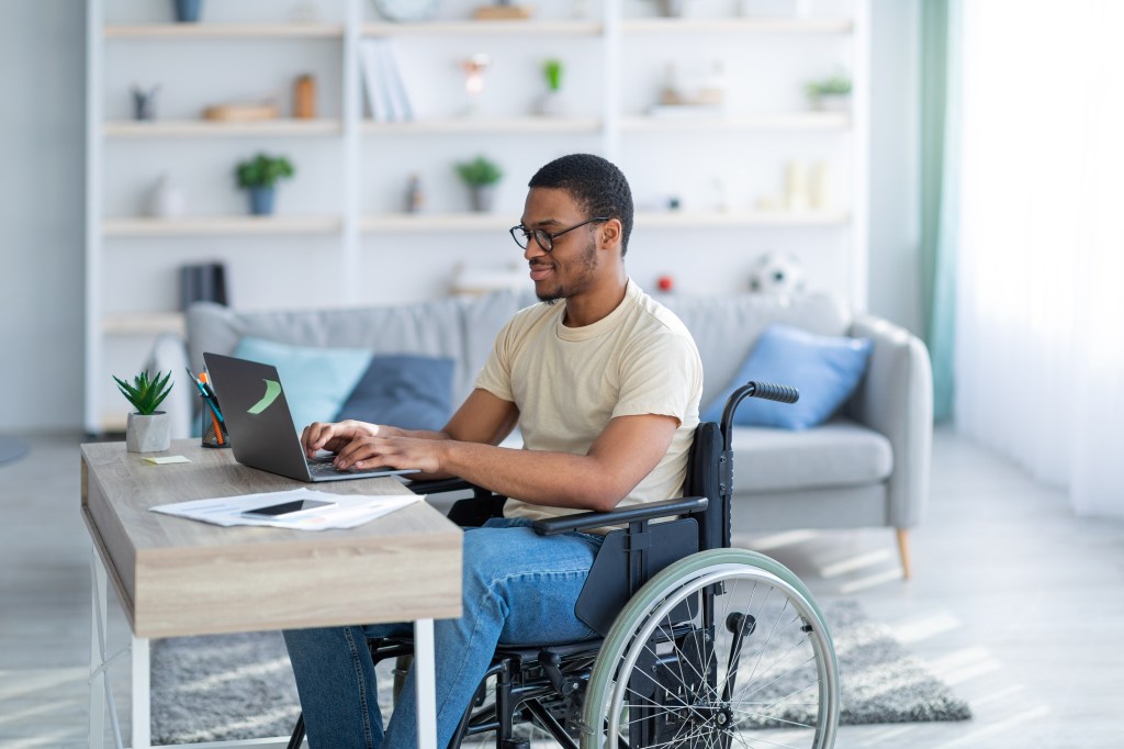 A black male in a wheelchair sits at an office desk and types on a laptop.
